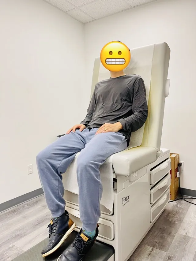 A man sitting on a padded exam chair in a US doctor's office, face covered with an emoji